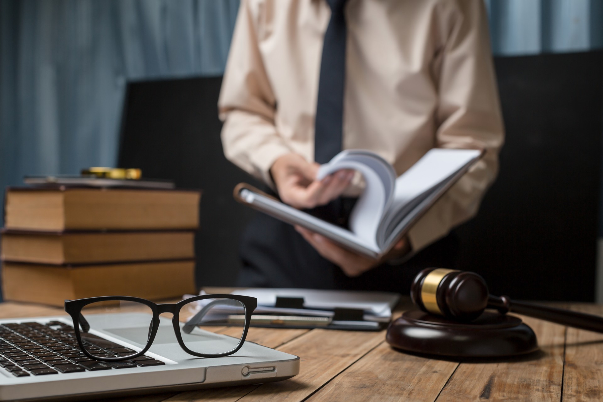 A close-up of a legal professional's desk featuring a gavel, law books, a laptop with glasses, and a person flipping through a book in the background.
