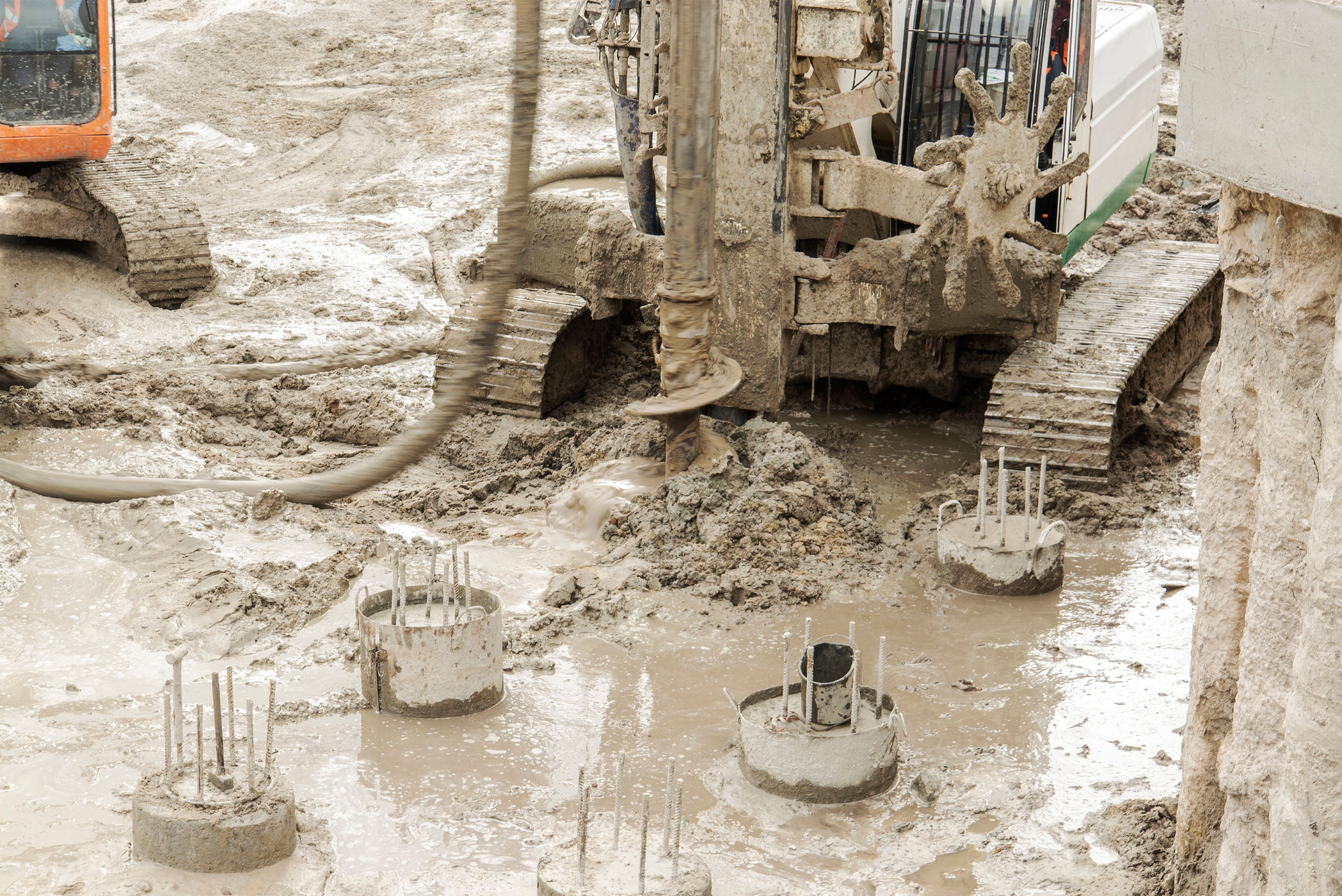 A heavy drilling rig operates in deep mud at a construction site, boring holes for deep foundations. Several newly completed circular concrete piles with exposed steel reinforcement bars are visible in the foreground.