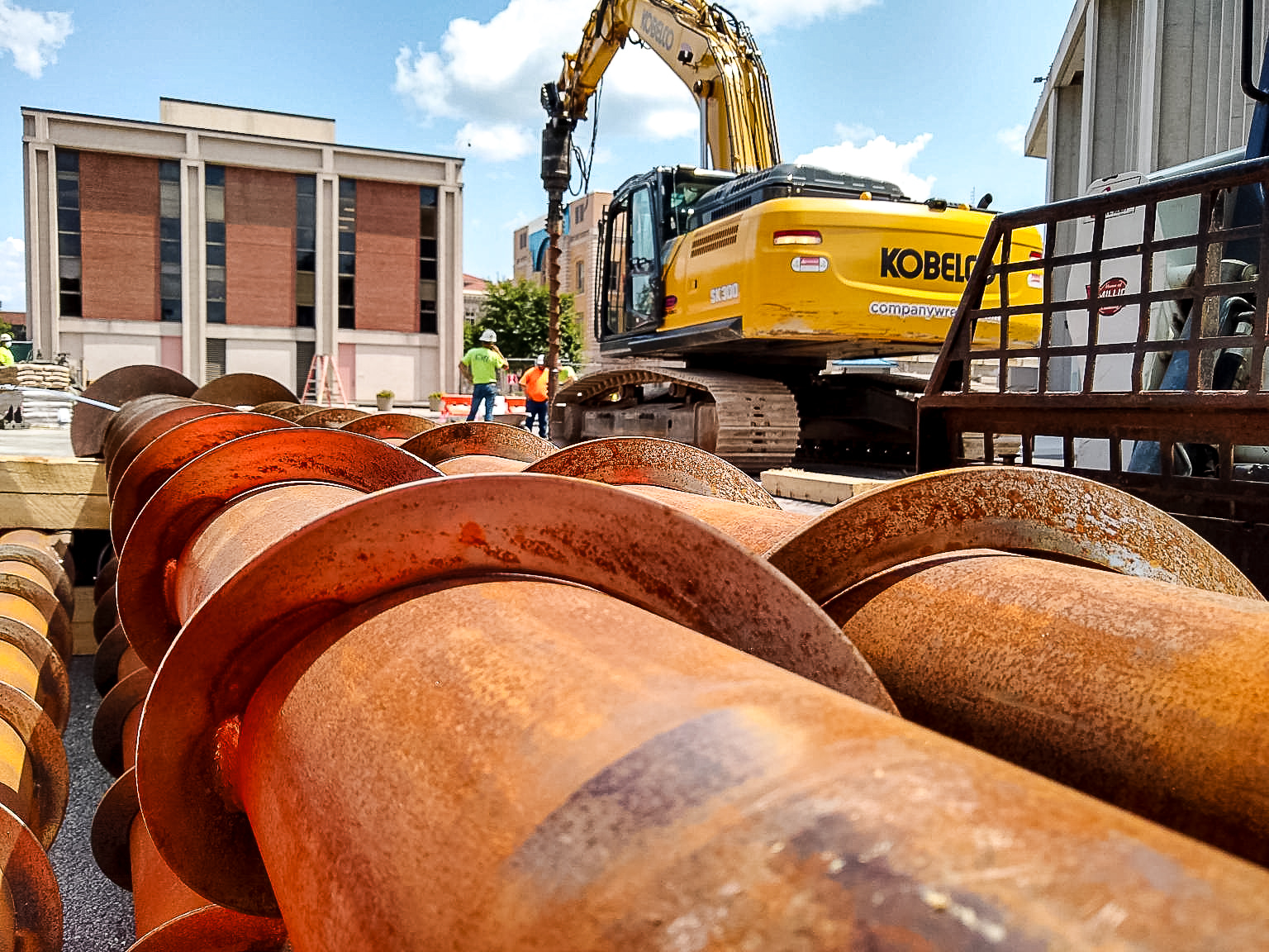 Close-up view of large, rusty helical auger pipes in the foreground, with a yellow Kobelco excavator and construction workers operating in the background on a job site.