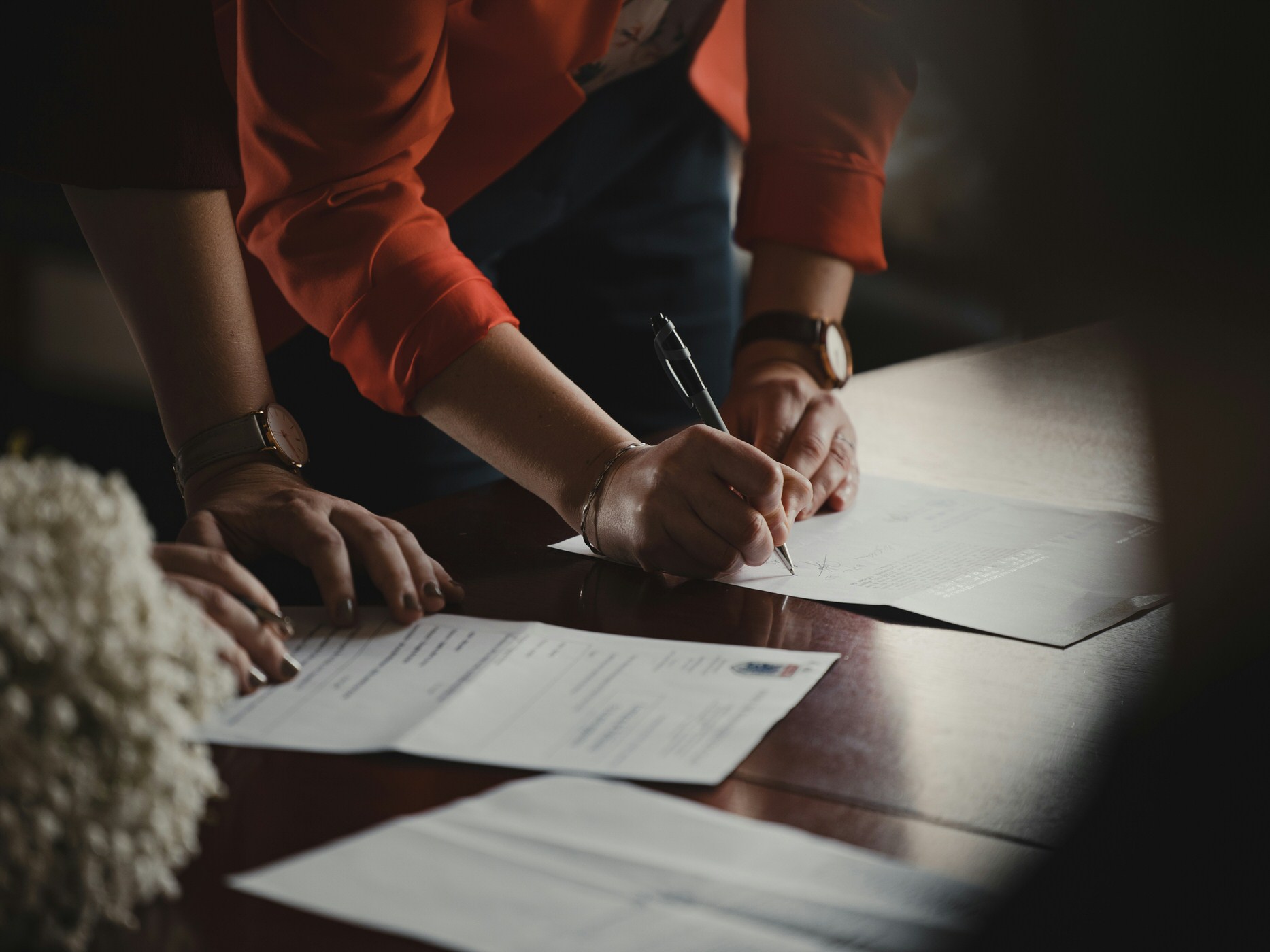 A person in an orange blazer signs a formal document with a pen, while another person's hands rest near a stack of papers on a wooden table.