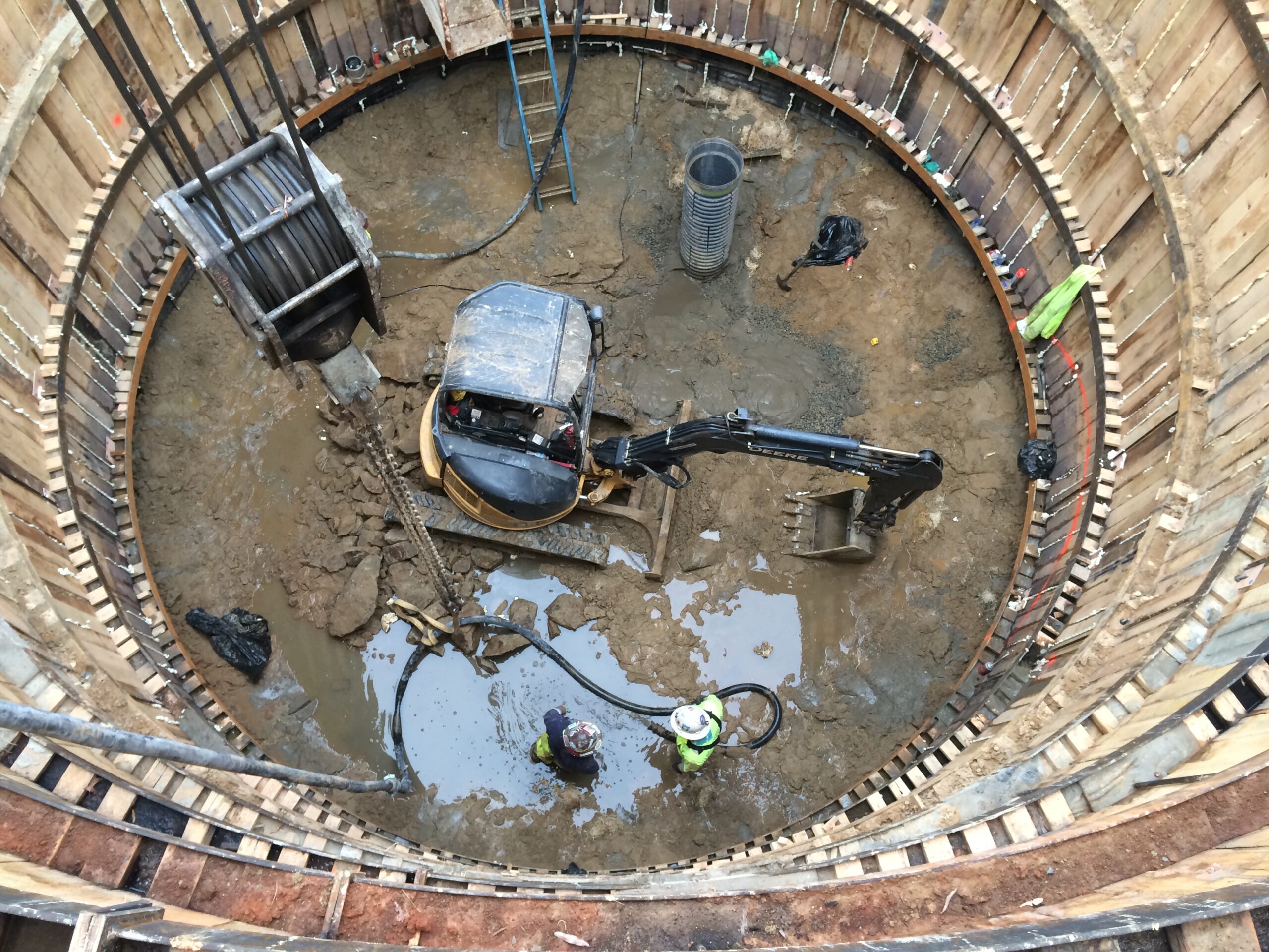 High-angle view of a circular construction shaft excavation, showing an excavator, two workers, mud, and timber/steel support rings.